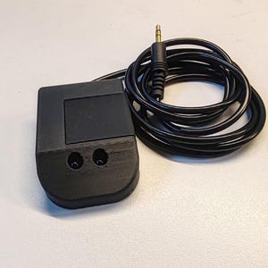 Può includere: Un dispositivo nero stampato in 3D con due piccoli fori e un cavo nero con un jack audio dorato.