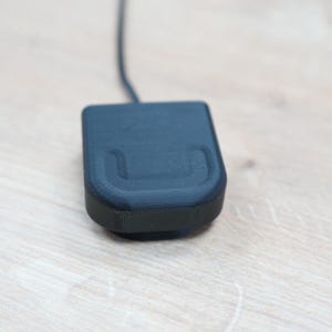 Può includere: Un caricabatterie nero rettangolare con un cavo attaccato. Il caricabatterie ha un'area curva e incassata nella parte superiore.