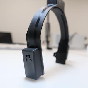 May include: A black, curved device with a rectangular component on one end. The device has a rounded arch shape with a textured section at the top. The background is a white surface with blurred objects.