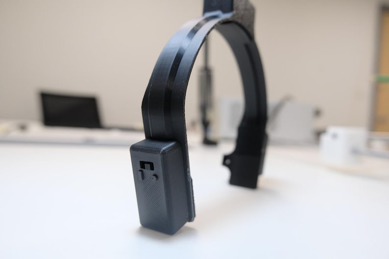 May include: A black, curved device with a rectangular component on one end. The device has a rounded arch shape with a textured section at the top. The background is a white surface with blurred objects.