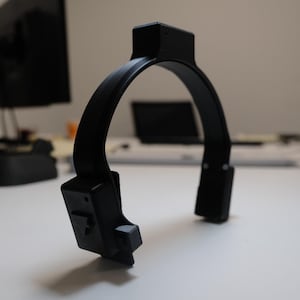 May include: A black, U-shaped device with a rectangular top and two rectangular extensions at the bottom. The device is made of a matte material and sits on a white surface. The background includes a computer monitor and other office equipment.