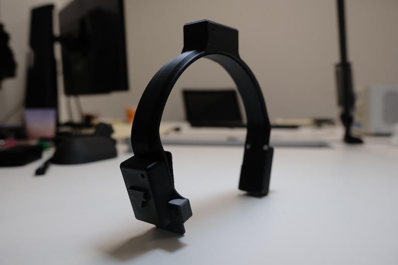 May include: A black, U-shaped device with a rectangular top and two rectangular extensions at the bottom. The device is made of a matte material and sits on a white surface. The background includes a computer monitor and other office equipment.