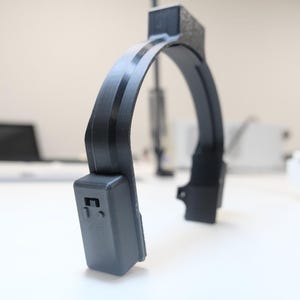 May include: A black, curved device with a rectangular base and a textured top. The device has a rounded arch with two dark stripes. The background is a white surface with blurred objects.