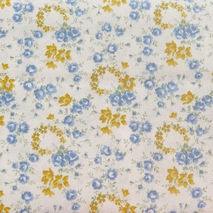 May include: Close-up of a white fabric with a floral pattern. The design features clusters of blue roses and yellow floral accents, with some arranged in circular patterns. The fabric has a delicate, vintage aesthetic.