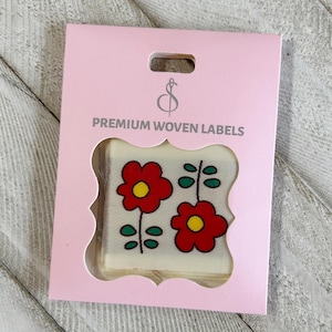 May include: A pink card with a die-cut window displaying four woven labels. Each label features two red flowers with yellow centers and green stems. The card reads "PREMIUM WOVEN LABELS" in black text.