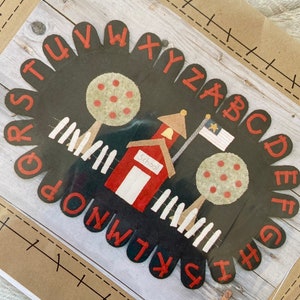 May include: A black oval-shaped schoolhouse design with red letters spelling out the alphabet. The design features a red schoolhouse with a white fence, two trees, and a flag. The word "School" is visible on the schoolhouse.