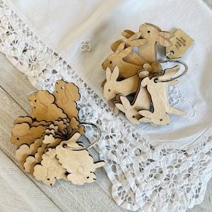 Könnte beinhalten: Ein Set aus Holz-Hasen- und Schaf-Anhängern mit einem silbernen Ring. Die Anhänger sind übereinander gestapelt und haben ein rustikales, natürliches Aussehen. Der Text auf dem Etikett lautet "Good morning bunny & sheep."