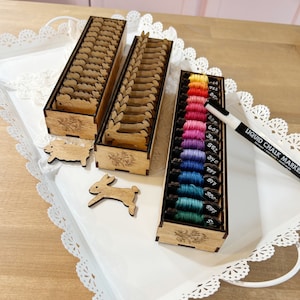 May include: Wooden storage box with multiple compartments for organizing embroidery floss. The box is filled with colorful floss spools labeled with numbers. A white liquid chalk marker is also included.