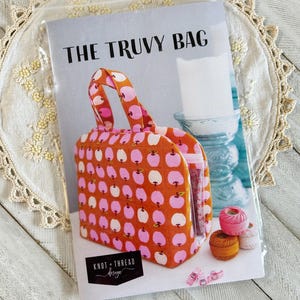 Pode incluir: Um padrão para "The Truvy Bag" com uma estampa colorida de maçãs. A bolsa tem uma alça e é feita de tecido laranja com desenhos de maçãs rosa e brancas. O texto "THE TRUVY BAG" é impresso na capa, junto com o logotipo "KNOT + THREAD design".