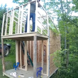 Puede incluir: Una estructura de madera de dos pisos en construcción, enmarcada con montantes y revestimiento de madera contrachapada. Una persona está de pie en el nivel superior, trabajando en el techo. La estructura está ubicada en una zona boscosa con un tractor visible en el fondo.