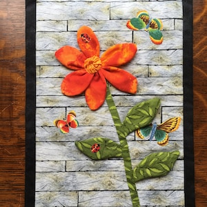 Könnte beinhalten: Ein handgefertigter Wandbehang mit einer leuchtend orangefarbenen Blume mit grünem Stiel und Blättern. Die Blume hat einen strukturierten Stoff und ist mit Marienkäfern verziert. Der Hintergrund ist ein graues Holzdielenmuster mit einem grünen Grasrand.