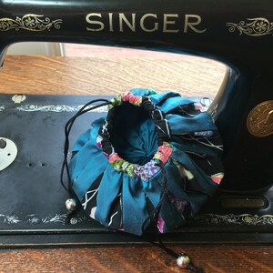May include: A teal blue fabric drawstring pouch with a floral print and black trim. The pouch is sitting on top of a vintage Singer sewing machine.