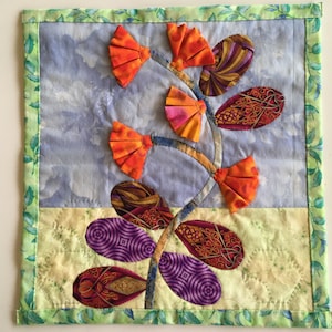 May include: A quilted wall hanging featuring a stylized flower with orange, purple, and burgundy fabric petals. The flower is set against a blue and white background with a yellow and white border.