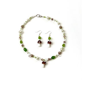 Pode incluir: Um conjunto de colar e brincos com tema de cogumelos. O colar e os brincos t&ecirc;m contas brancas, castanhas e verdes. Os brincos t&ecirc;m um berloque de cogumelo e berloques de folhas verdes. O colar tem um pingente de berloque de cogumelo.