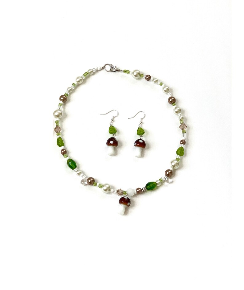 Pode incluir: Um conjunto de colar e brincos com tema de cogumelos. O colar e os brincos t&ecirc;m contas brancas, castanhas e verdes. Os brincos t&ecirc;m um berloque de cogumelo e berloques de folhas verdes. O colar tem um pingente de berloque de cogumelo.