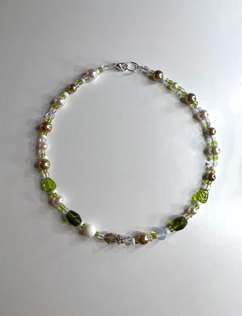 Pode incluir: Um colar de contas com contas brancas, castanhas e verdes. O colar inclui contas semelhantes a p&eacute;rolas, contas verdes facetadas e um pequeno pingente em forma de estrela. O colar &eacute; preso com um fecho prateado.