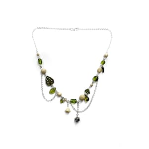 May include: A multi-strand silver-tone chain necklace featuring an assortment of olive green leaf-shaped beads, round cream-colored beads, and small dangling charms. The design incorporates layered chains that create a draped effect across the front.