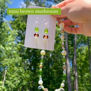 Pode incluir: Um conjunto de joias com tema de cogumelos. O colar e os brincos apresentam pingentes de cogumelos marrons e brancos, contas de folhas verdes e contas transparentes. O texto "mini brown mushroom" &eacute; vis&iacute;vel.