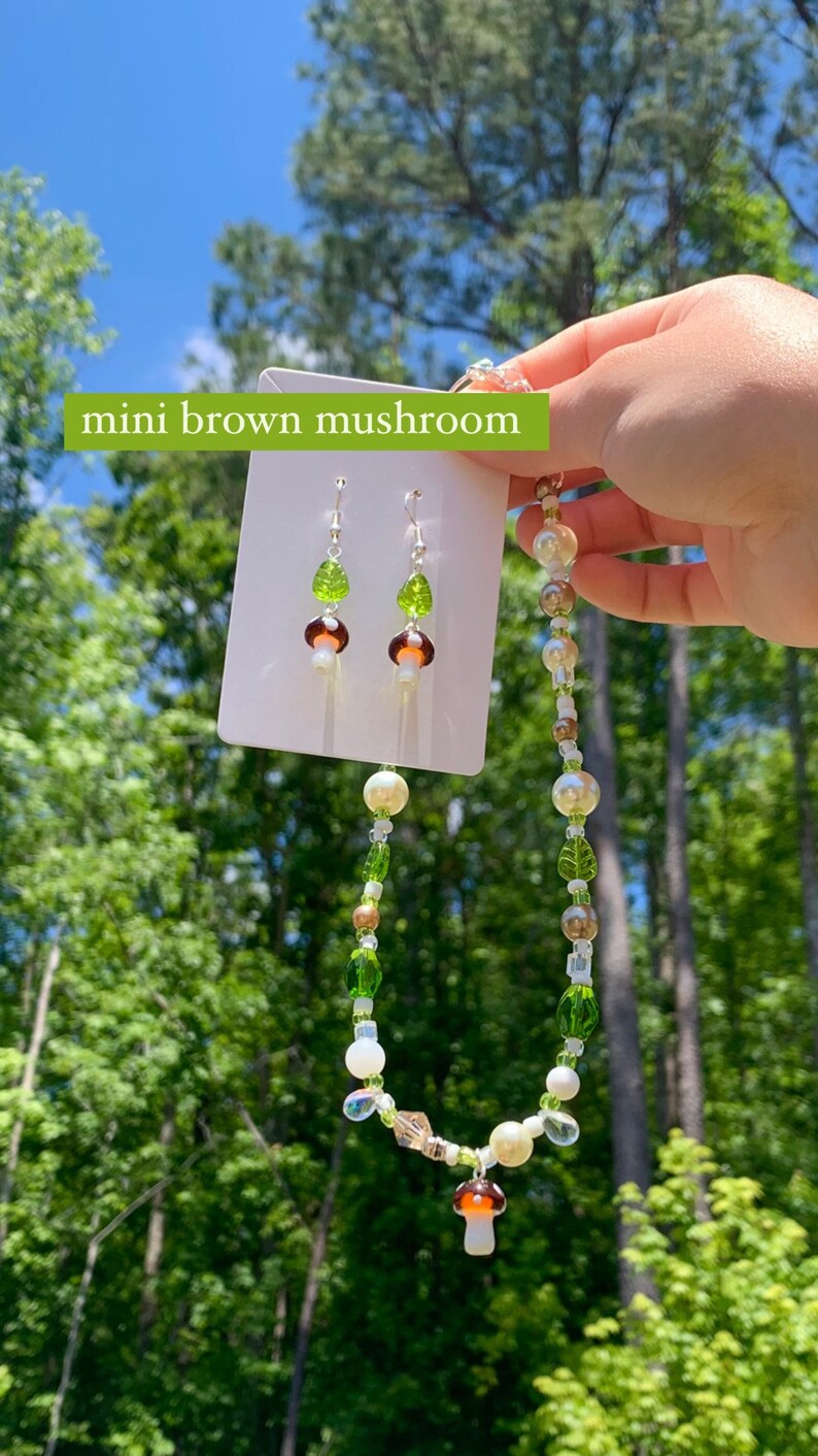 Pode incluir: Um conjunto de joias com tema de cogumelos. O colar e os brincos apresentam pingentes de cogumelos marrons e brancos, contas de folhas verdes e contas transparentes. O texto "mini brown mushroom" &eacute; vis&iacute;vel.