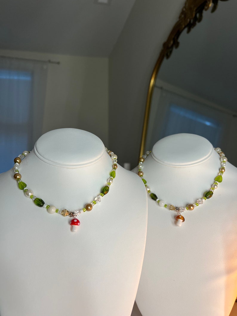 Pode incluir: Dois colares de contas com pingentes de cogumelos. Os colares apresentam uma mistura de contas verdes, brancas, douradas e transparentes. Cada colar tem um pequeno pingente de cogumelo com uma tampa vermelha e um caule branco. Os colares s&atilde;o exibidos em bustos brancos.