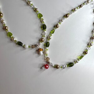 Pode incluir: Dois colares de contas com um design caprichoso. Os colares apresentam uma mistura de contas brancas, douradas e verdes, com detalhes em forma de folha. Pequenos pingentes de cogumelos com tampas vermelhas e brancas pendem dos colares.