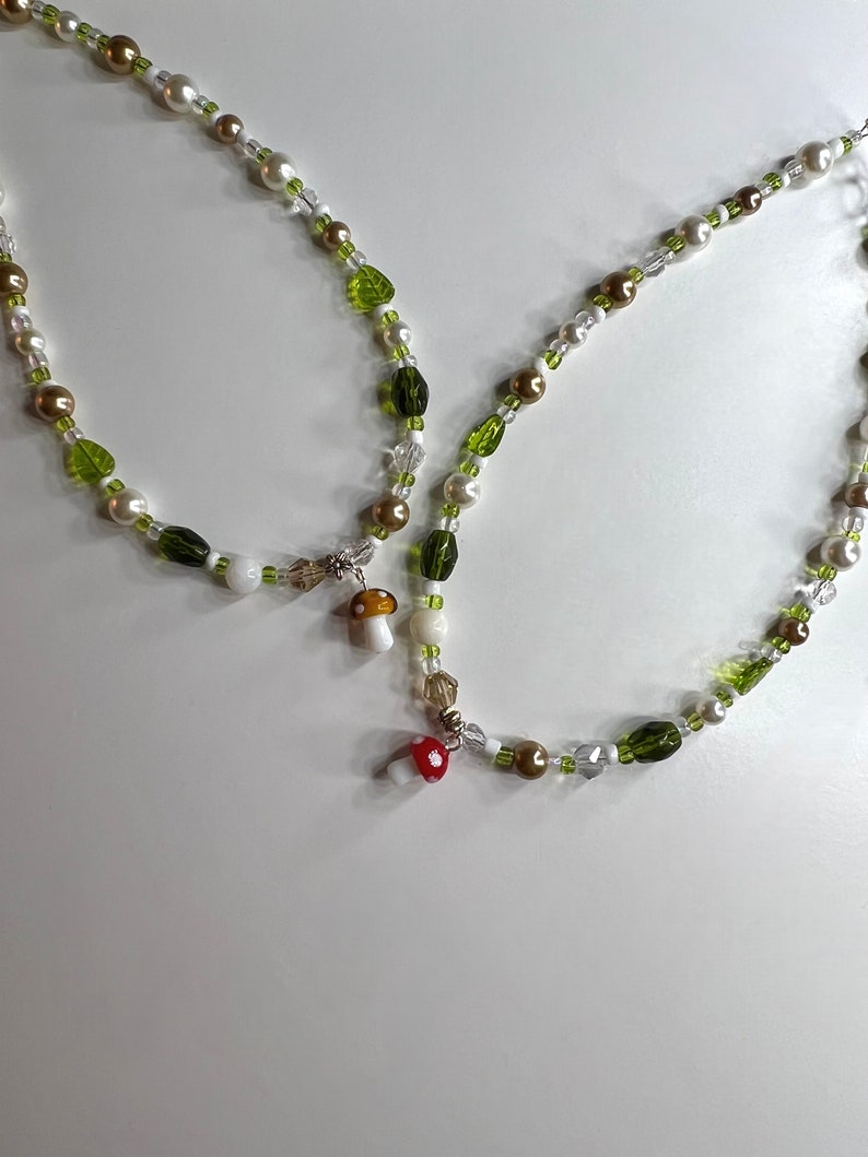 Pode incluir: Dois colares de contas com um design caprichoso. Os colares apresentam uma mistura de contas brancas, douradas e verdes, com detalhes em forma de folha. Pequenos pingentes de cogumelos com tampas vermelhas e brancas pendem dos colares.
