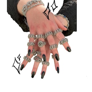 May include: Silver-toned chainmail rings and bracelets. The rings feature intricate woven designs, with some showcasing a central gemstone. The jewelry is displayed on a hand with black nail polish, creating a striking contrast.