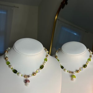 Pode incluir: Duas gargantilhas de contas expostas em bustos brancos. As gargantilhas apresentam uma mistura de contas brancas, verdes e castanhas, com um pingente de cogumelo. Um pingente de cogumelo &eacute; branco e verde, o outro &eacute; branco e roxo.