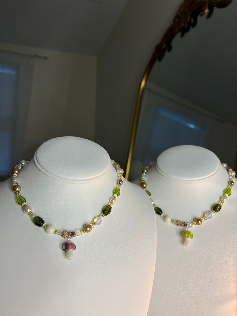 Pode incluir: Duas gargantilhas de contas expostas em bustos brancos. As gargantilhas apresentam uma mistura de contas brancas, verdes e castanhas, com um pingente de cogumelo. Um pingente de cogumelo &eacute; branco e verde, o outro &eacute; branco e roxo.