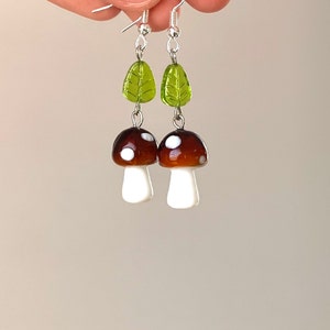 Pode incluir: Um par de brincos de cogumelo feitos &agrave; m&atilde;o. Cada brinco apresenta um cogumelo castanho com manchas brancas e um caule branco, ligado a uma conta de folha verde. Os brincos penduram-se de ganchos prateados.