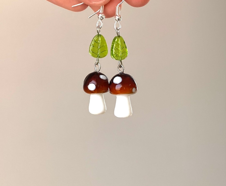 Pode incluir: Um par de brincos de cogumelo feitos &agrave; m&atilde;o. Cada brinco apresenta um cogumelo castanho com manchas brancas e um caule branco, ligado a uma conta de folha verde. Os brincos penduram-se de ganchos prateados.