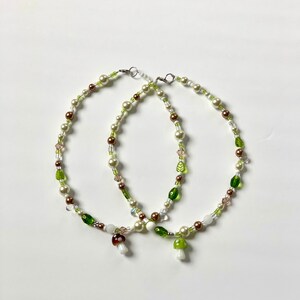Pode incluir: Dois colares de contas com contas brancas, castanhas e verdes. Cada colar tem um pingente de cogumelo. Os colares est&atilde;o em um fundo branco.