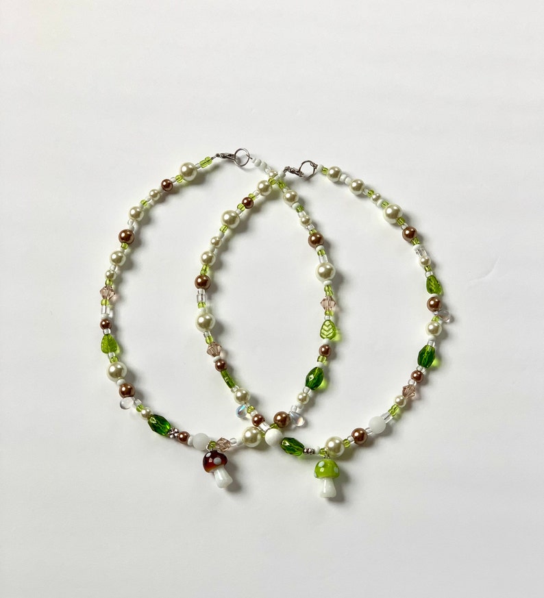 Pode incluir: Dois colares de contas com contas brancas, castanhas e verdes. Cada colar tem um pingente de cogumelo. Os colares est&atilde;o em um fundo branco.