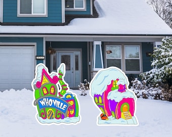 May include: Two colorful, whimsical yard signs depicting cartoon houses covered in snow. One sign reads "Whoville" in blue lettering. The houses are in shades of pink, green, yellow, and purple, with whimsical architectural details.