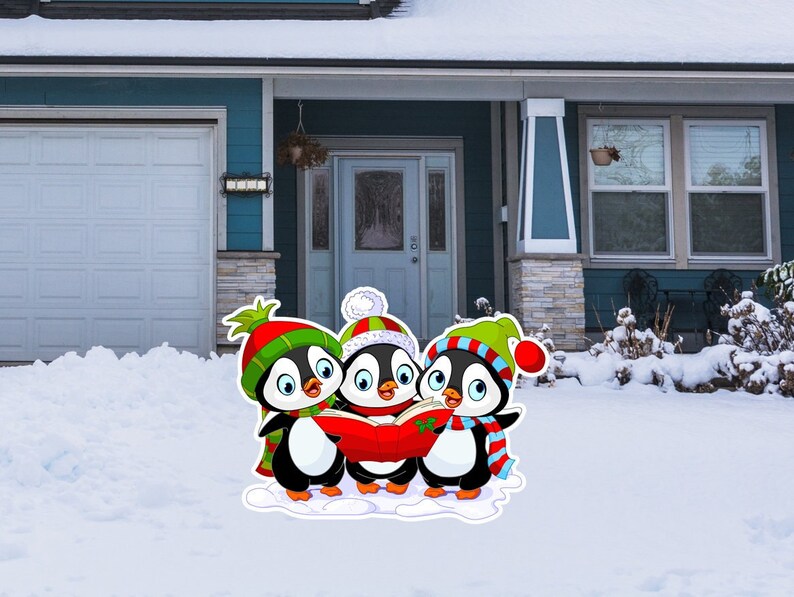 Op de afbeelding: Een kleurrijke kerstversiering voor buiten met drie zingende cartoonpingu&iuml;ns. De pingu&iuml;ns dragen rode, groene en witte mutsen en sjaals en houden een rood zangboek vast. De decoratie staat in een besneeuwde tuin voor een huis.