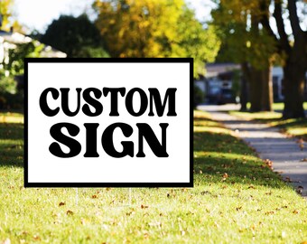 May include: A white sign with the words "CUSTOM SIGN" in bold, black letters. The sign is displayed on a grassy lawn with a residential neighborhood in the background. The sign is framed with a black border.