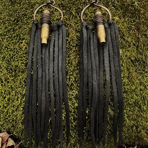 May include: A pair of handmade earrings featuring a brass bullet casing and long, black leather fringe. The earrings have a copper-colored hoop and are set against a mossy green background.
