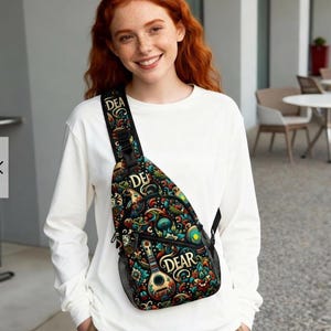 May include: A black sling bag with a colorful, ornate design featuring musical instruments and the word "DEAR". The bag has a mesh side pocket and a black strap. The bag is worn over the shoulder.