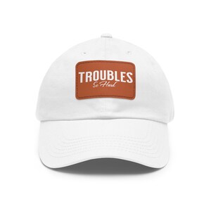 May include: White baseball cap with a brown leather patch that says "TROUBLES So Hard" in white text.