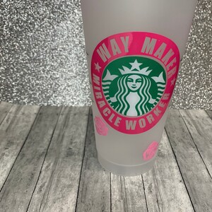 以下が含まれることがあります： 白い再利用可能なカップで、ピンクの円形のデザインが施され、「ウェイメーカーミラクルワーカー」と書かれており、中央に緑色のスターバックスのロゴがあります。カップの底にはピンクのバラが飾られています。