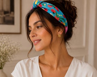May include: A bright blue fabric headband featuring a knotted top detail and a colorful abstract floral pattern in shades of red, yellow, and green. The headband has a smooth finish and is worn across the crown of the head, framing the hair.