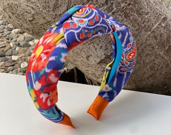 May include: A colorful knotted headband with a vibrant floral and paisley pattern in shades of blue, red, yellow, and orange. The headband has orange tips and a turquoise accent.