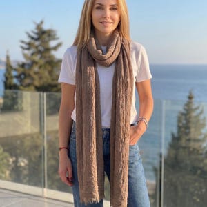 May include: A long, rectangular, taupe-colored scarf with a crinkled, textured fabric finish and frayed edges. The scarf is draped loosely around the neck, hanging down the front of a plain white short-sleeved t-shirt and blue denim jeans.
