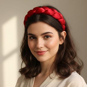 May include: A braided red satin headband. The headband is a vibrant, solid colour and is worn by a person with dark wavy hair. The person is wearing a light-coloured top. The background is a neutral colour.