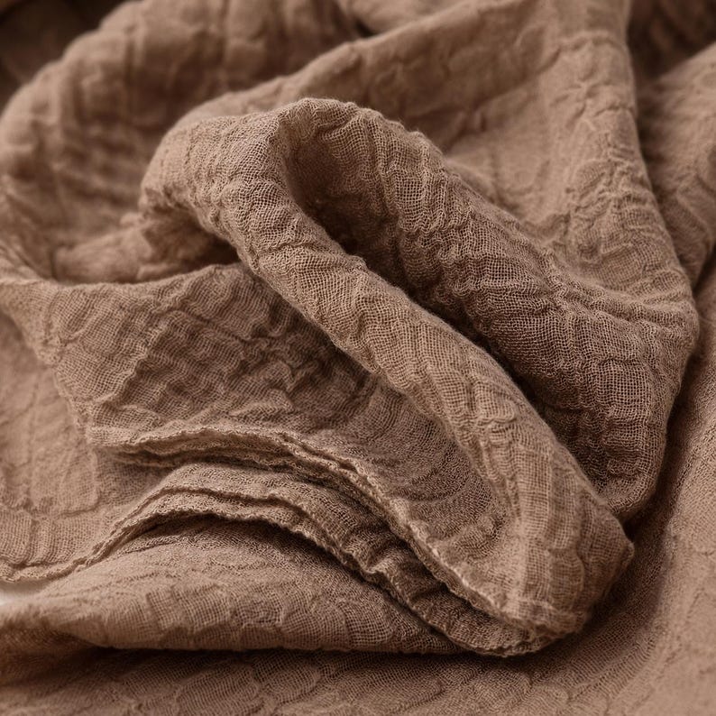 May include: A light brown fabric with a distinct crinkled, textured surface. The material features a loose, open weave construction with visible folds and drapes that create a layered appearance across the entire surface of the textile.