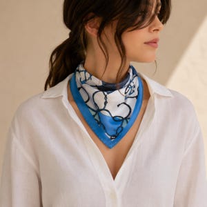 Peut inclure: Un foulard carré en soie présentant un fond blanc avec un motif de chaînes bleu foncé et noir, bordé d'une bordure bleu uni. Le foulard est plié en triangle et noué autour du cou, porté avec une chemise blanche unie à boutons.