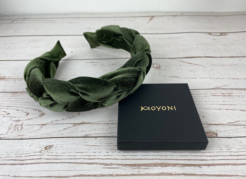 May include: A dark green velvet headband with a braided design. The headband is on a wooden surface next to a black box with the word "XOYONI" printed in gold.