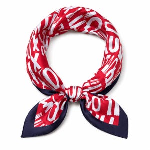May include: A square scarf featuring a vibrant red background with a repeating white brushstroke letter pattern. The scarf is tied in a knot at the centre, revealing a solid dark navy blue border along the bottom edges of the fabric material.