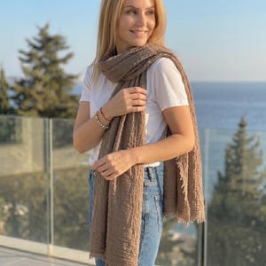 May include: A long, rectangular, light brown scarf with a crinkled, textured fabric surface and frayed edges. The scarf is draped over the shoulders and hangs down the front, worn over a plain white t-shirt and blue denim jeans.