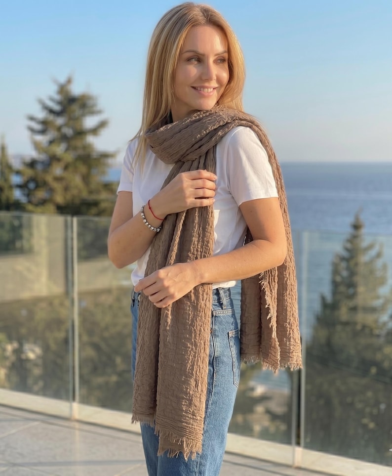 May include: A long, rectangular, light brown scarf with a crinkled, textured fabric surface and frayed edges. The scarf is draped over the shoulders and hangs down the front, worn over a plain white t-shirt and blue denim jeans.